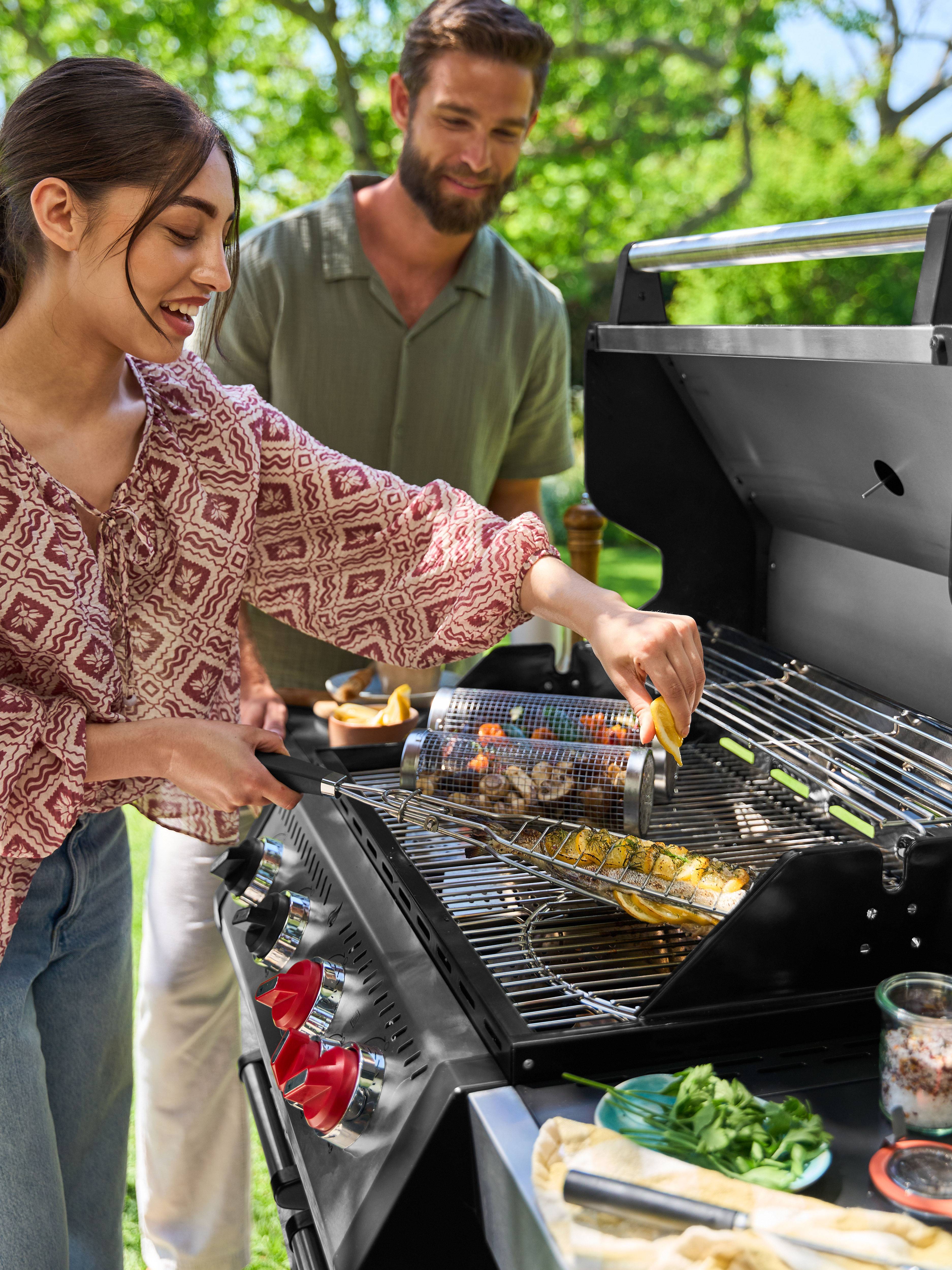 Smiling woman grilling fish and vegetables on a gas barbecue, man watching from behind.