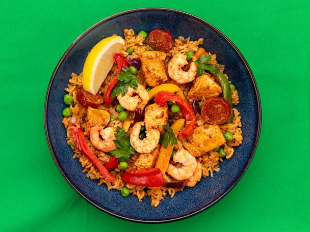 A blue plate of paella with chicken, prawns, chorizo, peppers, peas, and a lemon wedge.