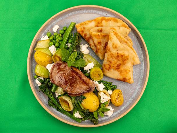 Lamb steak with roasted potatoes, green vegetables, feta, mint, and flatbreads on a plate.