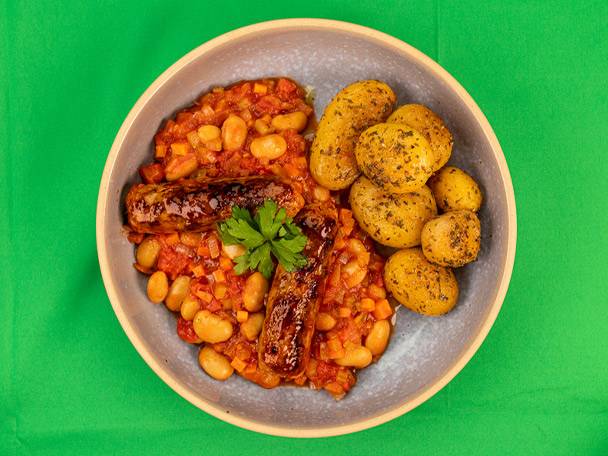 Sausages with baked beans and roasted potatoes in a bowl on a green background.