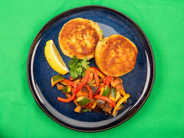 Two golden-brown fishcakes with roasted vegetables and a lemon wedge on a blue plate.