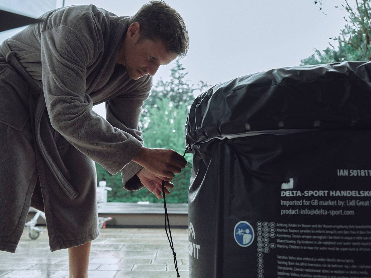 A man in a bathrobe adjusts the cover of a Delta-Sport hot tub.
