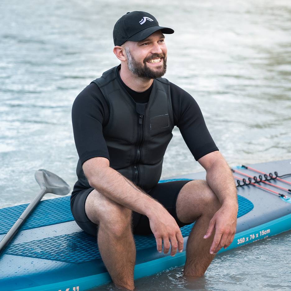 Smiling man in a life vest and cap, sitting on a paddleboard in the water.