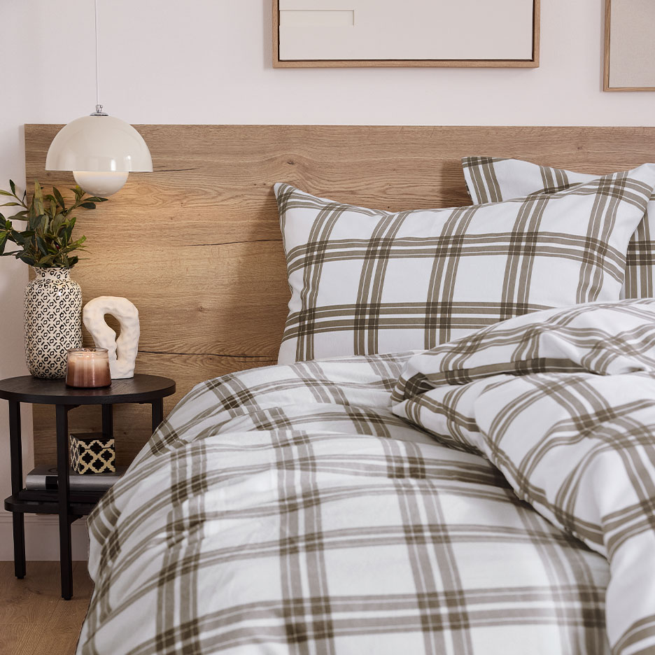 Bed with wooden headboard and plaid bedding, with a nightstand and lamp.