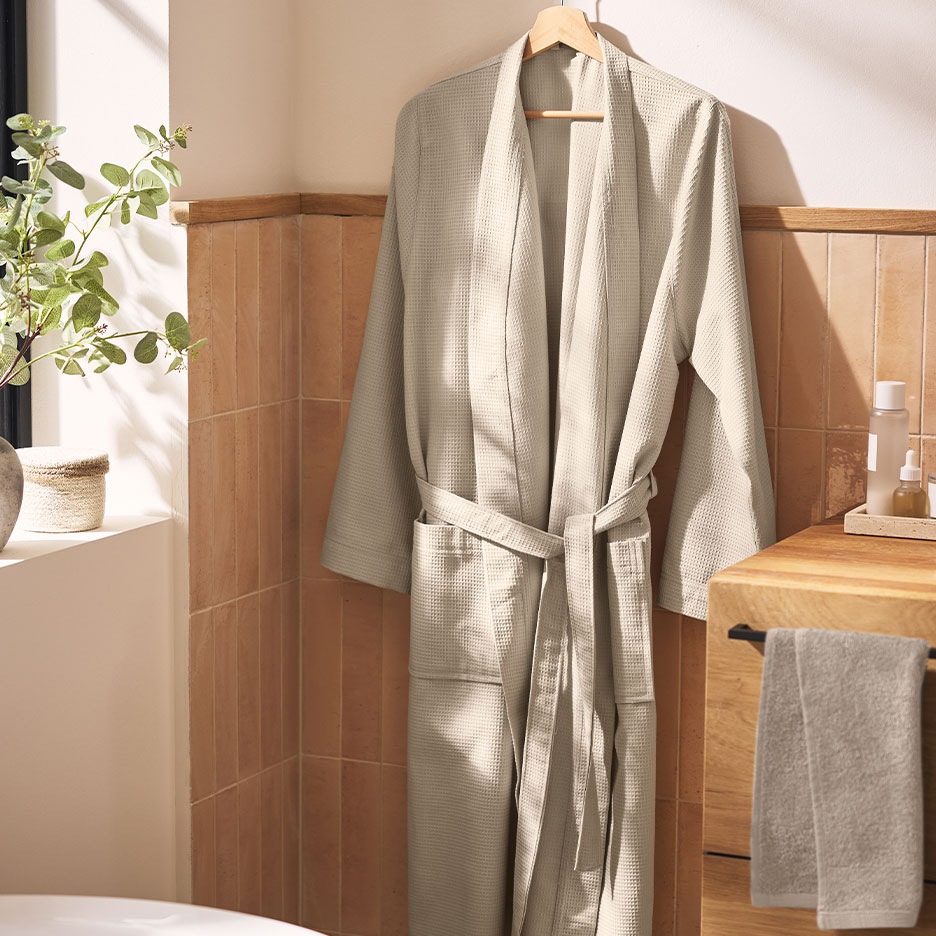 Beige waffle bathrobe hanging in a bathroom with brown tiles and plants.
