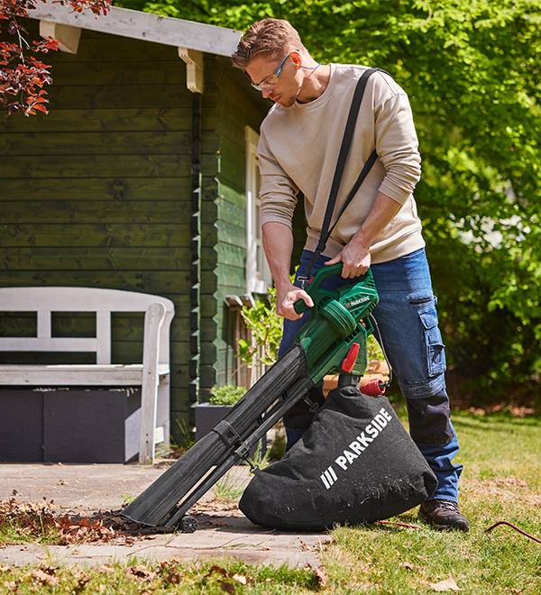 A man uses a Parkside leaf blower and collection bag to clean leaves in his garden.