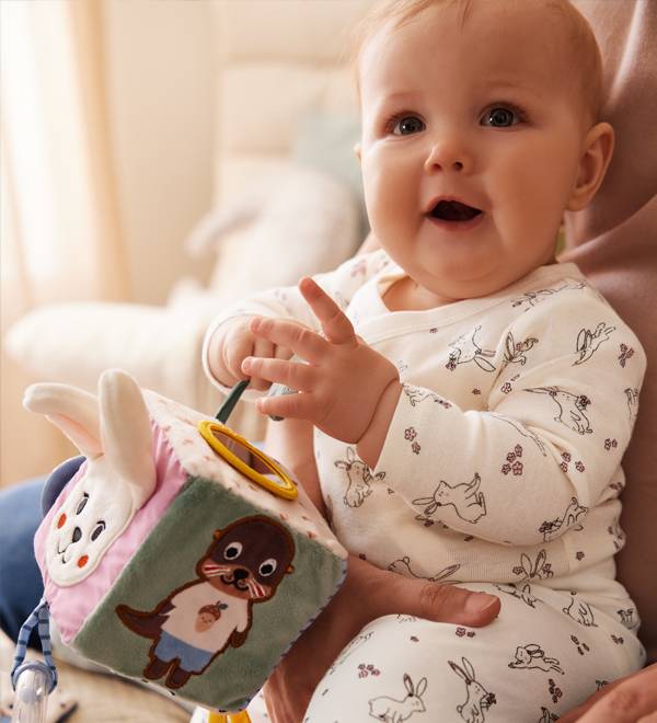 A baby in bunny-print pajamas holds a soft activity cube with animal designs.