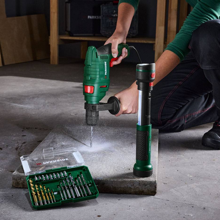 Person drilling a hole in a slab with a drill, next to a flashlight and a drill bit set.