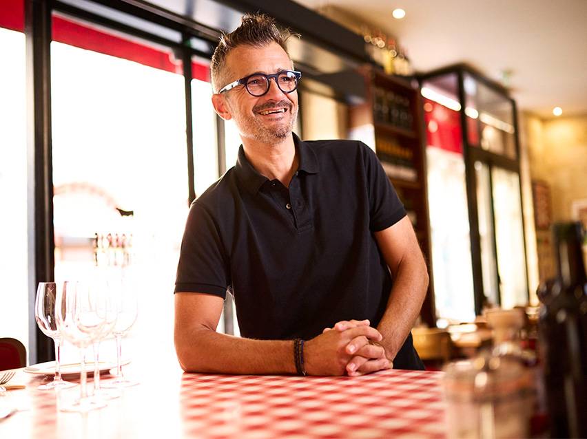Smiling man in black polo shirt, leaning on a red and white checkered table in a restaurant.
