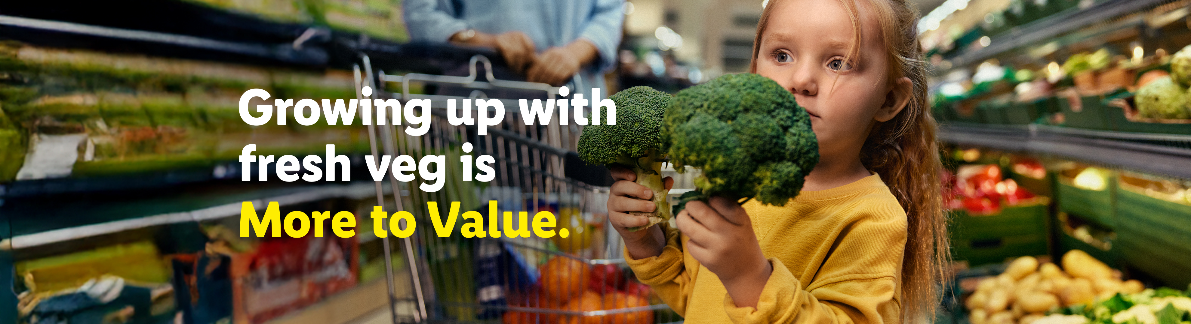 A child holding two broccoli heads in a grocery store with a shopping cart and fresh produce.