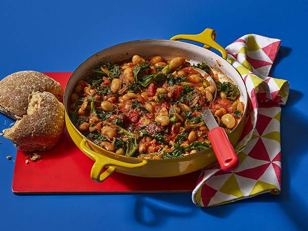 Hearty bean and kale stew in a yellow pot with bread rolls on a red board.