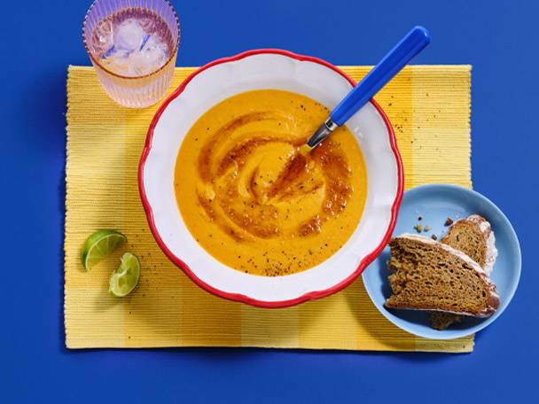 A bowl of orange soup with a spoon, a glass of water, and bread on a blue background.