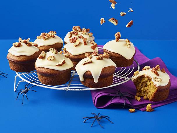 Pumpkin spice muffins with cream cheese frosting and pecans, some falling, on a blue background.