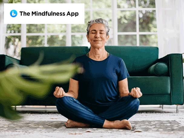 Woman meditating on a rug with a green sofa and 'The Mindfulness App' logo.