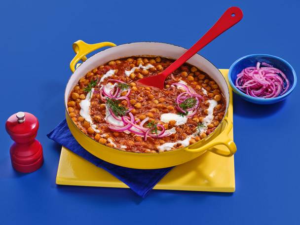 A yellow pot of chickpea curry with a red spoon, red pepper grinder, and a blue bowl of pickled onions.