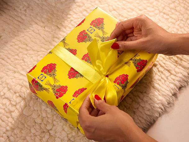 Hands tying a yellow ribbon on a gift box with red floral and Lidl logo wrapping paper.