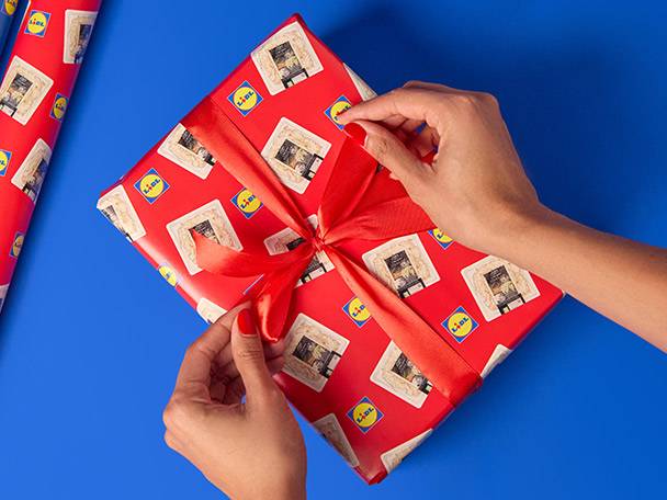 Hands tying a red ribbon on a gift wrapped in red paper with Lidl logos and cheese.