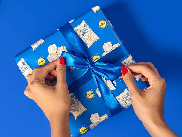 Hands tying a blue ribbon on a gift wrapped in Lidl ice cube paper.