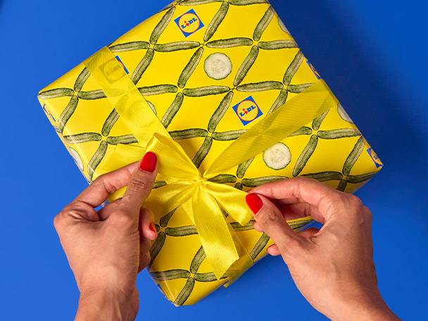 Hands tying a yellow ribbon on a Lidl-branded gift wrapped in cucumber-patterned paper.