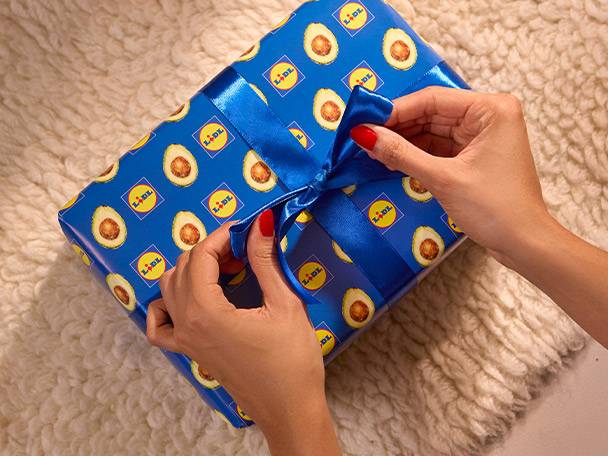 Hands tying a blue ribbon on a Lidl avocado-patterned gift box on a white rug.