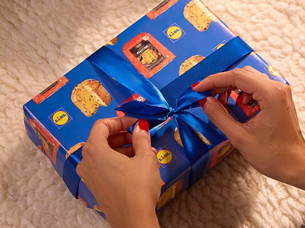 Hands tying a blue ribbon on a gift box wrapped in Lidl panettone-themed paper.