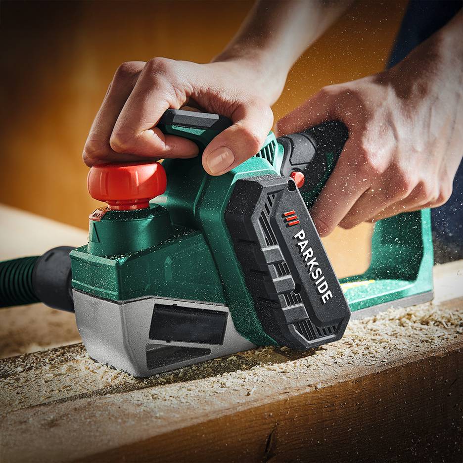 Hands using a green Parkside electric planer on a wooden board, creating wood shavings.