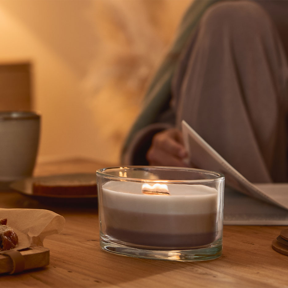 Burning three-layered candle in a glass, with a person reading in the background.