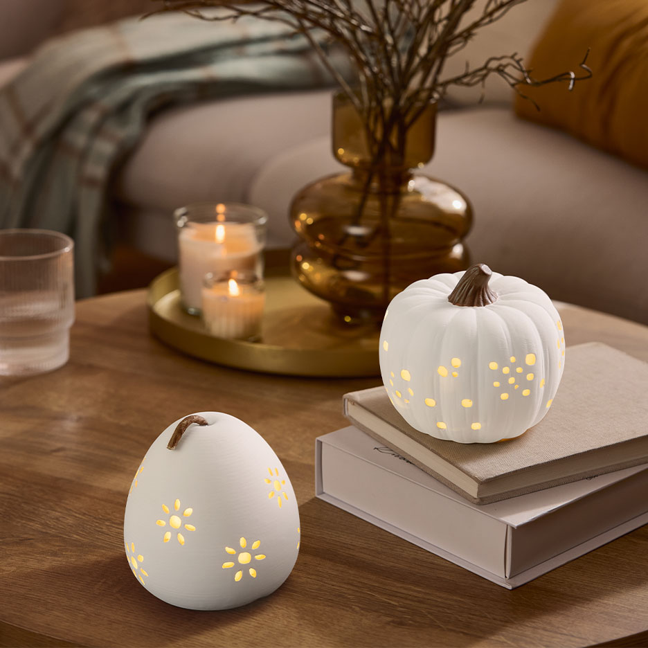 Two white illuminated decorative pumpkins on a wooden table with candles and a vase.