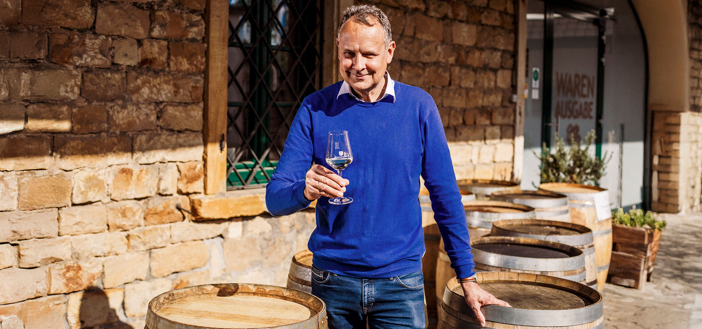Smiling man in blue sweater holding a glass of white wine in front of a stone wall.