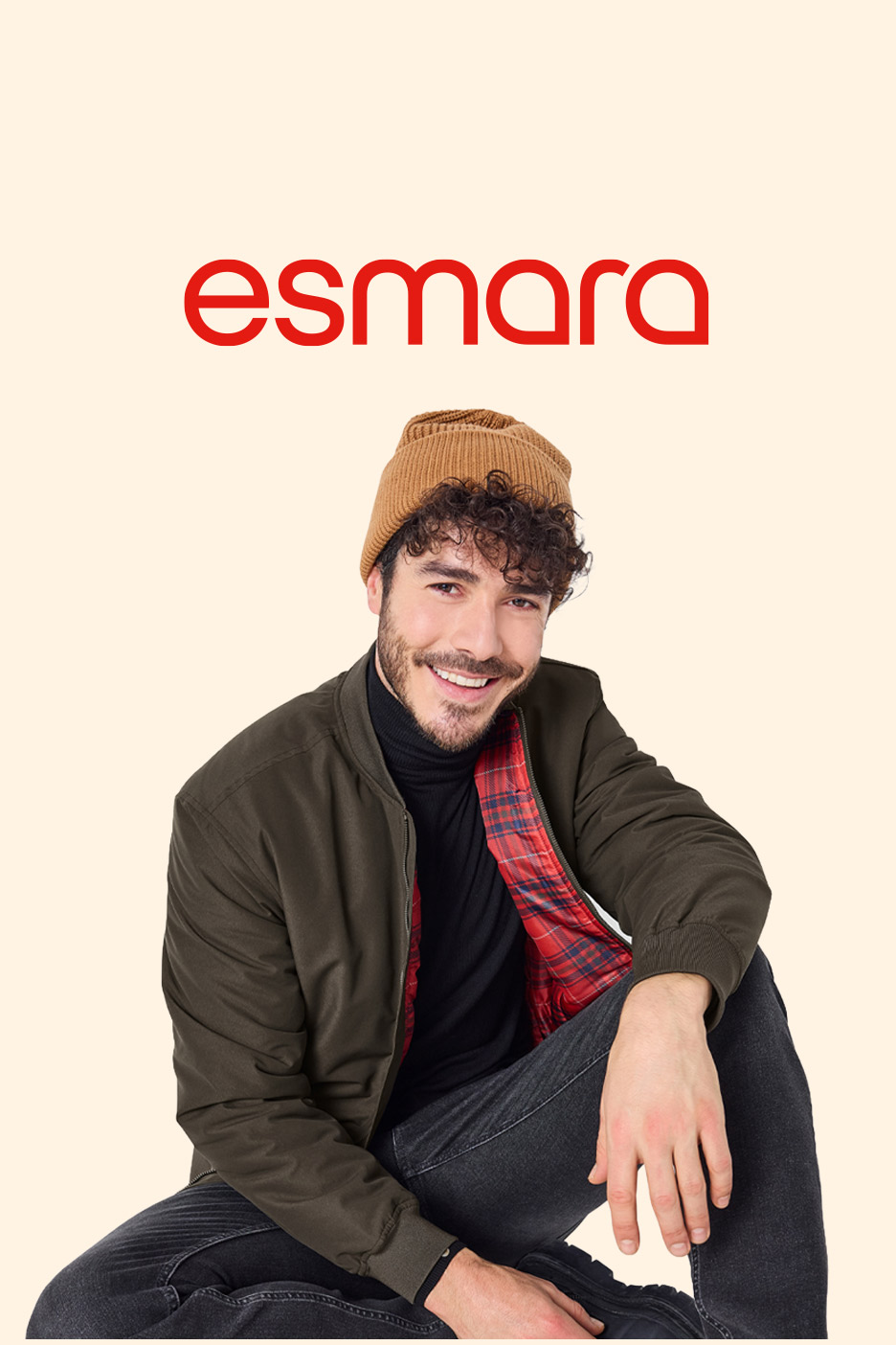 Smiling man in a brown beanie, dark green bomber jacket with red plaid lining, and black jeans.