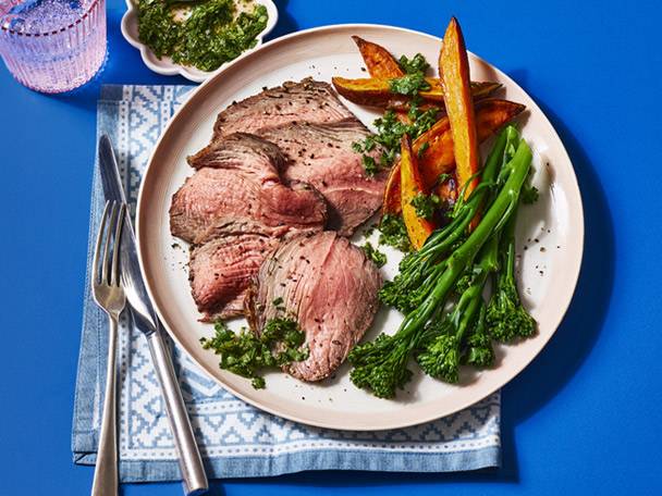 Sliced roast beef with sweet potato wedges, broccolini, and green sauce on a plate.