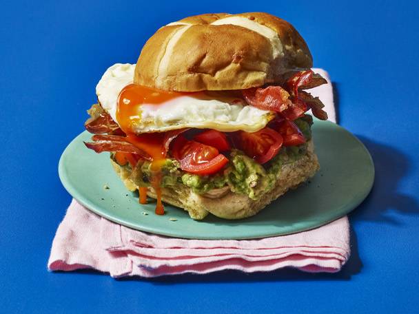 Hot cross bun burger with egg, bacon, avocado, and tomatoes on a blue background.