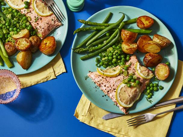 Two plates of salmon with roasted potatoes, green beans, peas, and lemon slices.