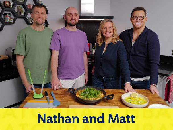 Four people in a kitchen with a pan of food and a bowl of salad on a wooden counter.