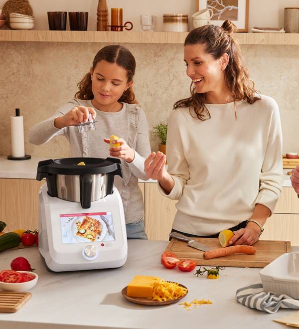 A mother and daughter cooking together with a white kitchen appliance.