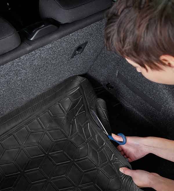 A person cuts a black rubber car trunk mat to fit the vehicle's space.