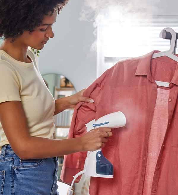 A woman uses a handheld garment steamer to refresh a coral-colored shirt hanging on a clothes rack.