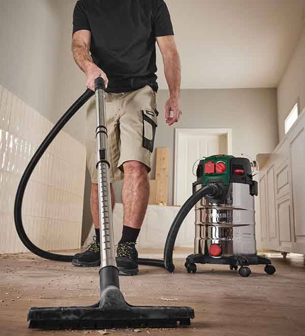 A person uses a Bosch industrial vacuum cleaner to clean up sawdust on a wood floor.