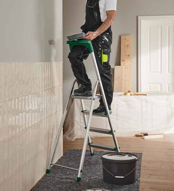 Man on a step ladder applying plaster to a wall, with a bucket nearby.