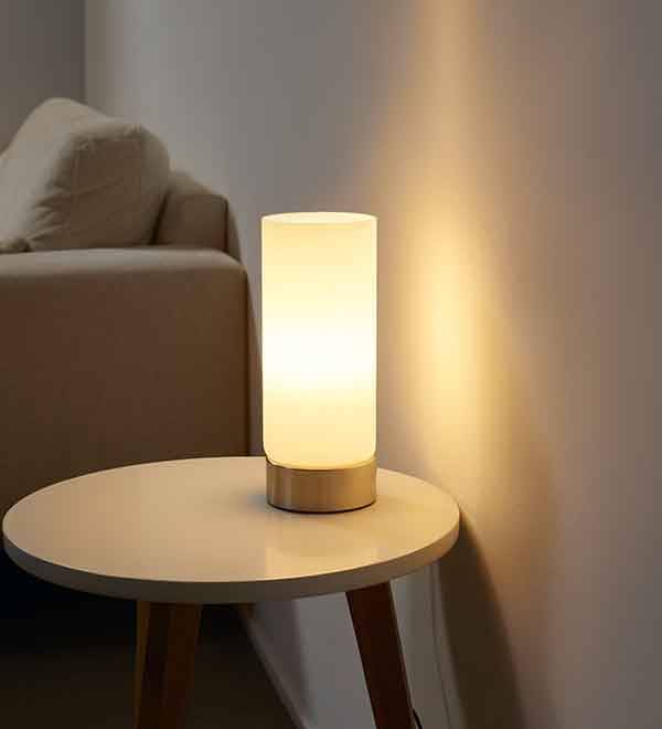 A cylindrical table lamp with a glowing white shade on a round side table.