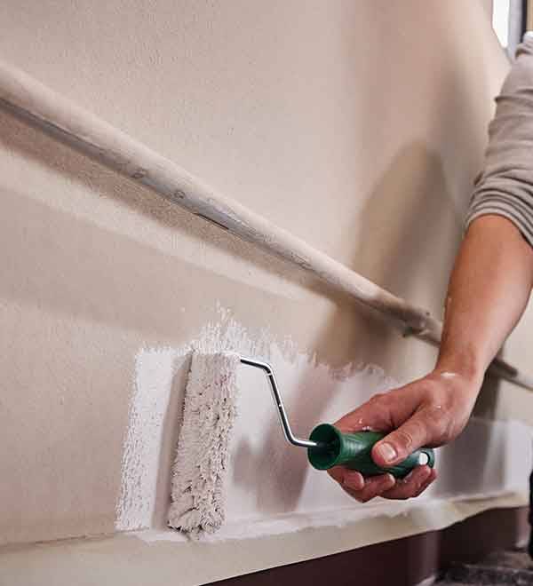 A person uses a paint roller to apply white paint to a beige wall.