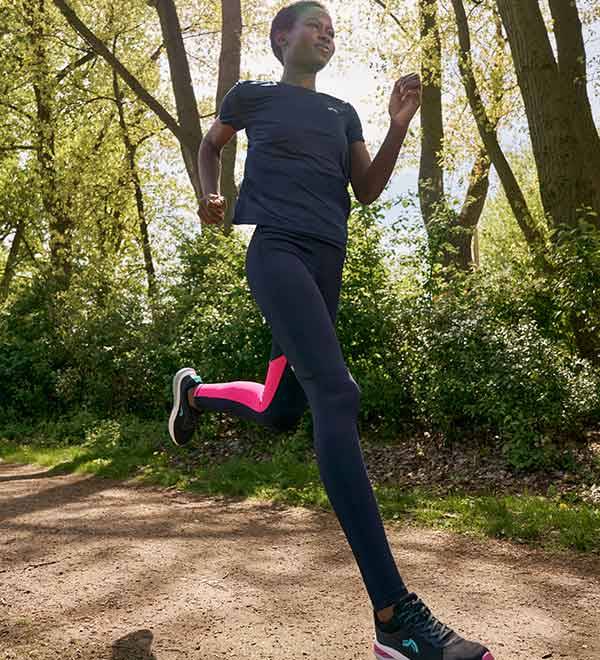 A woman in a black t-shirt and black leggings with pink accents runs on a dirt path.
