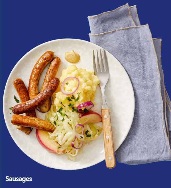 A plate of sausages with mashed potatoes, cabbage, and apple slices, next to a fork and napkin.