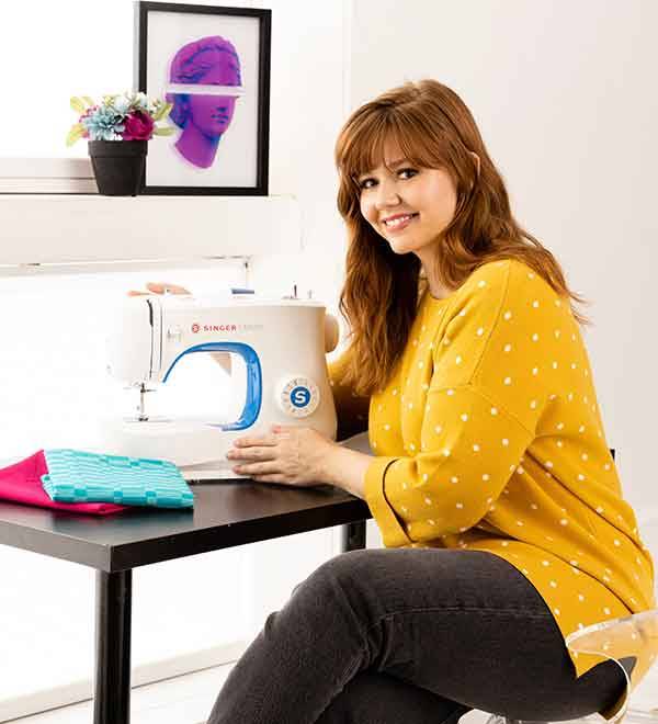 A woman sits at a table using a Singer sewing machine.