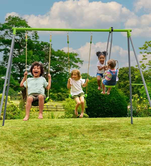 Three children enjoy swinging on a TP Toys metal swing set in a lush green backyard.