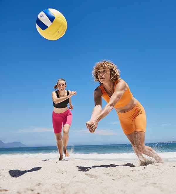 Two women play volleyball on a beach, one in pink shorts and the other in orange shorts.