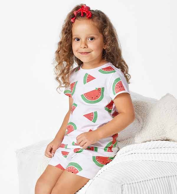 A girl in a watermelon-print pajama set sits on a white bed.