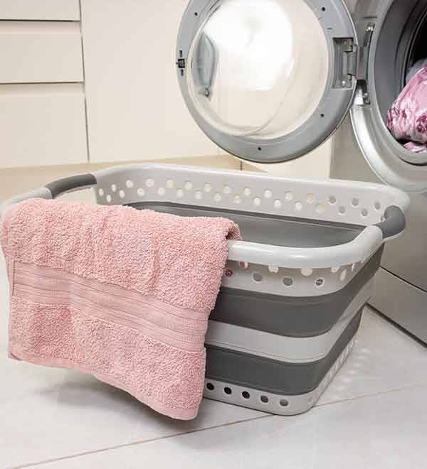 A collapsible laundry basket sits next to a washing machine, holding a pink towel.