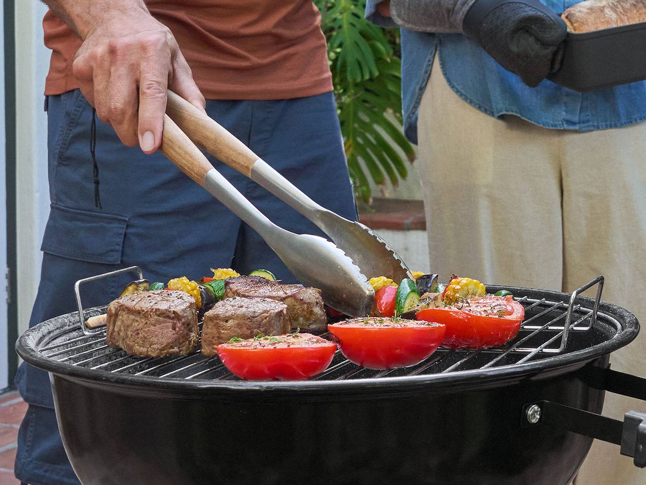 Man grilling meat, vegetables, and tomatoes on a barbecue, with a woman holding bread.