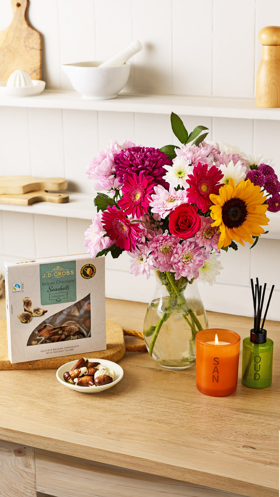 A bouquet of colorful flowers, chocolate seashells, and scented candles on a kitchen counter.